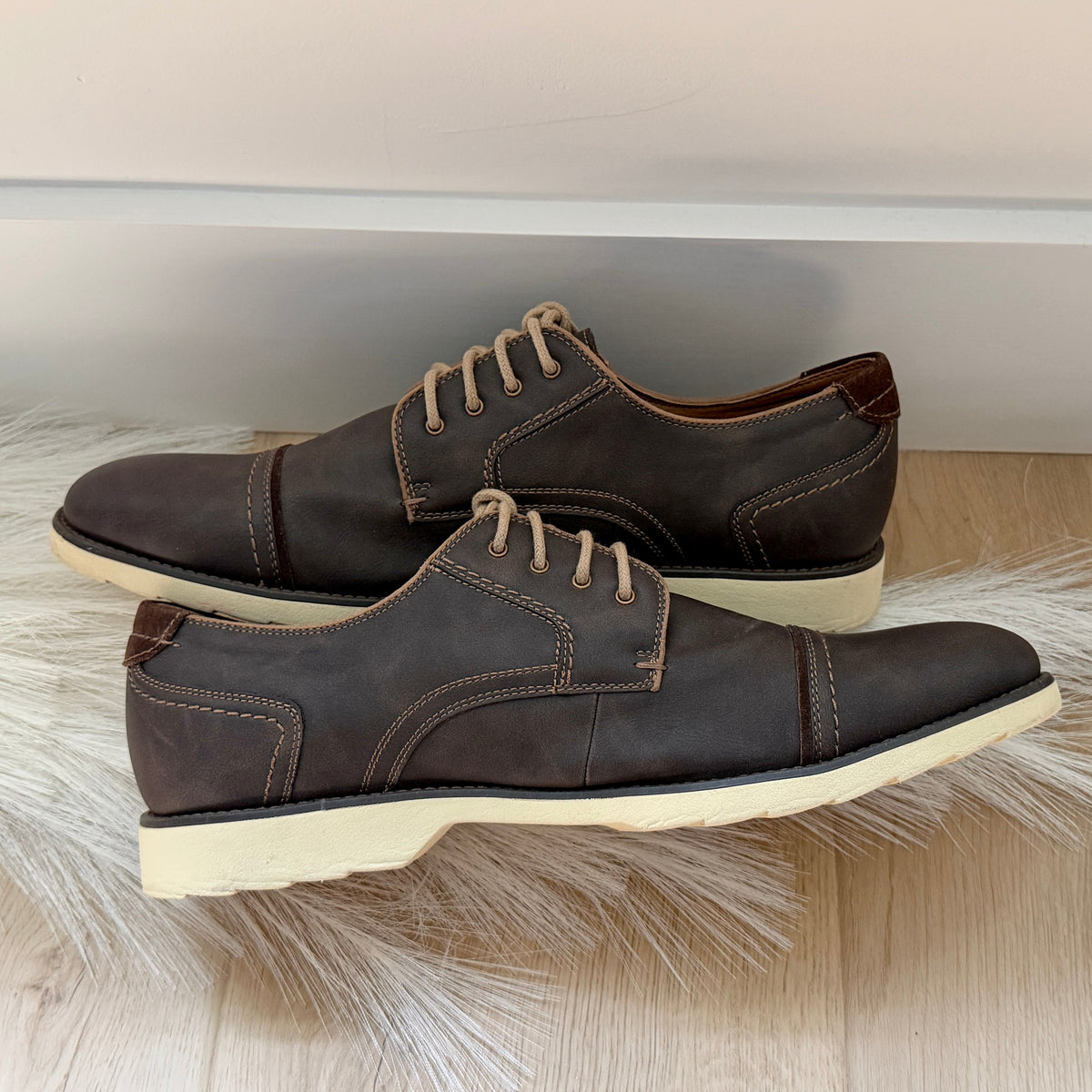 A pair of dark brown leather cap-toe oxford shoes with beige laces and off-white soles, displayed on a white faux fur rug over a light wood floor.